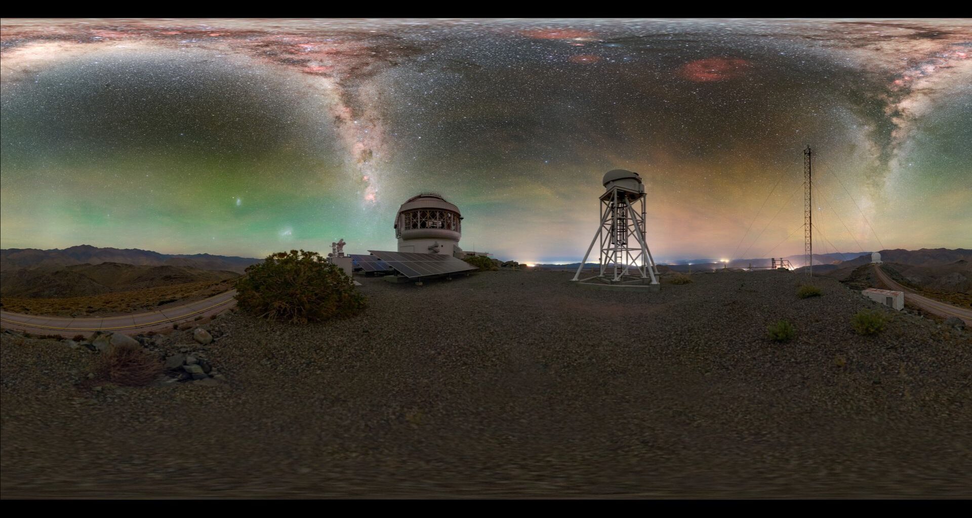 A glowing panorama shows jagged shapes of blue and purple of the Milky Way in a green and yellow night sky. Below are metal domed buildings with a barren landscape around them