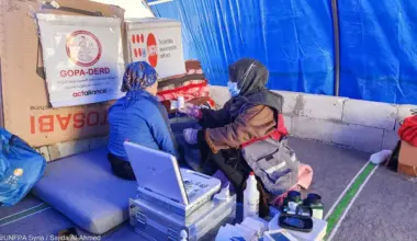 Inside a blue tarpaulin tent, a female health worker is seen from the side sitting in front of a female patient who is on a mattress. Beside them are medical equipment, stacked blankets and boxes with the UNFPA logo