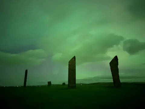 Lisa MacKenzie Tall standing stones rise from a dark, grassy landscape under a glowing green sky. Clouds drift across the scene, giving it an eerie, twilight atmosphere.