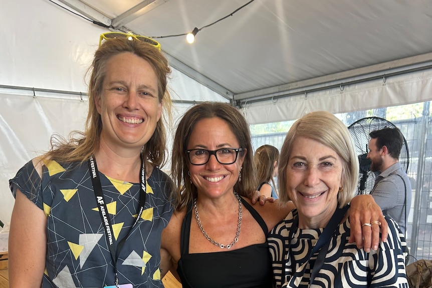 Three women, Anna Clark, 48, Clare Wright, 56, and Louise Adler, 71, in lanyards, pose smiling together in a tent.
