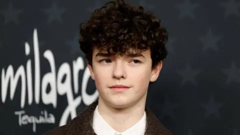 Reuters Owen Cooper, who is wearing a brown leather jacket and white shirt, is standing on the red carpet at the 31st Annual Critics Choice Awards in Santa Monica, California.