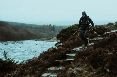 Clorroe Cam/Montane Spine Race A lone runner dressed in dark green makes her way down steps next to a large river with a forest in the distance