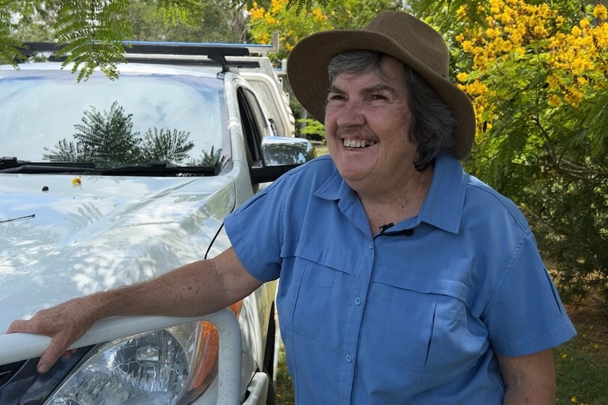 A woman in a blue shirt and hat smiles while leaning against a white car.