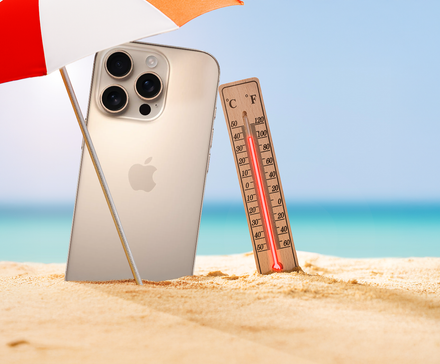 An iPhone and a thermometer in the sand under a beach umbrella, with the ocean in the background.