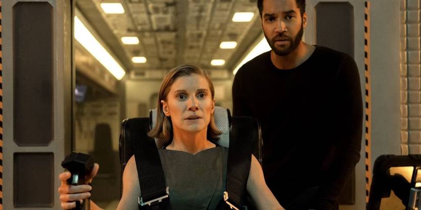 Katee Sackhoff strapped into a spacecraft chair grips a control handle while a man stands behind her in a futuristic cabin.