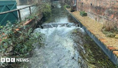 Donald Trump and Keir Starmer sit in a formal drawing room setting  on arm chairs with US and UK  flags behind them. Both have serious expressions and are looking out and not at each other.