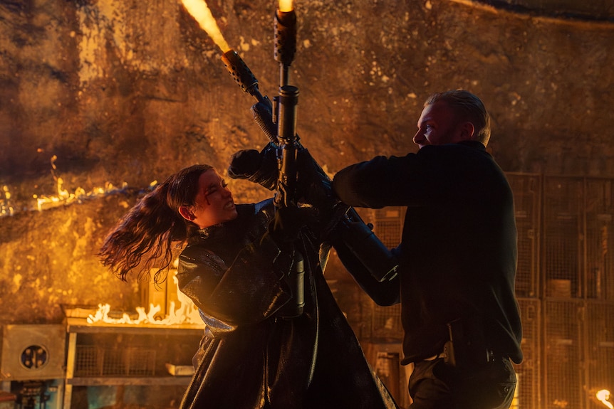Ana de Armas holding a flame thrower wrestles with a man who is also holding a flamethrower.