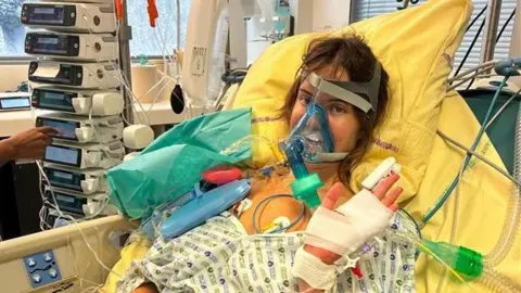 Faye Greenwood A woman with brown hair in a hospital bed with multiple tubes attached to her body.