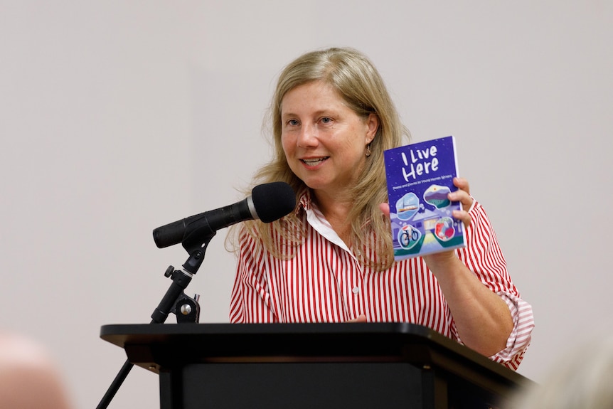 Rosemarie Milsom, a middle-aged woman, stands behind a lectern, holding a children's book that reads "I live here".