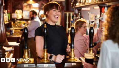 Woman with  black, shoulder-length hair, wearing a light blue shirt and brown apron pulls a pint of lager behind the bar in a pub