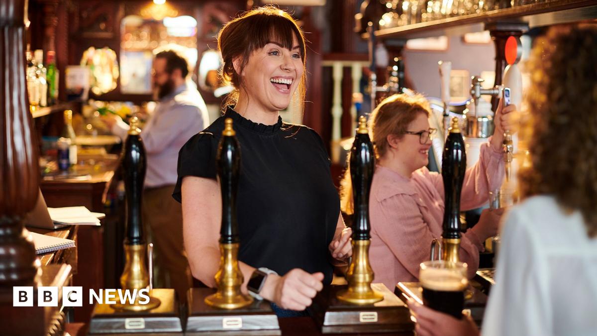 Woman with  black, shoulder-length hair, wearing a light blue shirt and brown apron pulls a pint of lager behind the bar in a pub