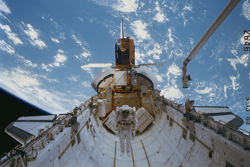 An astronaut floating in the open belly of a space shuttle with a satellite attached above him and Earth behind.