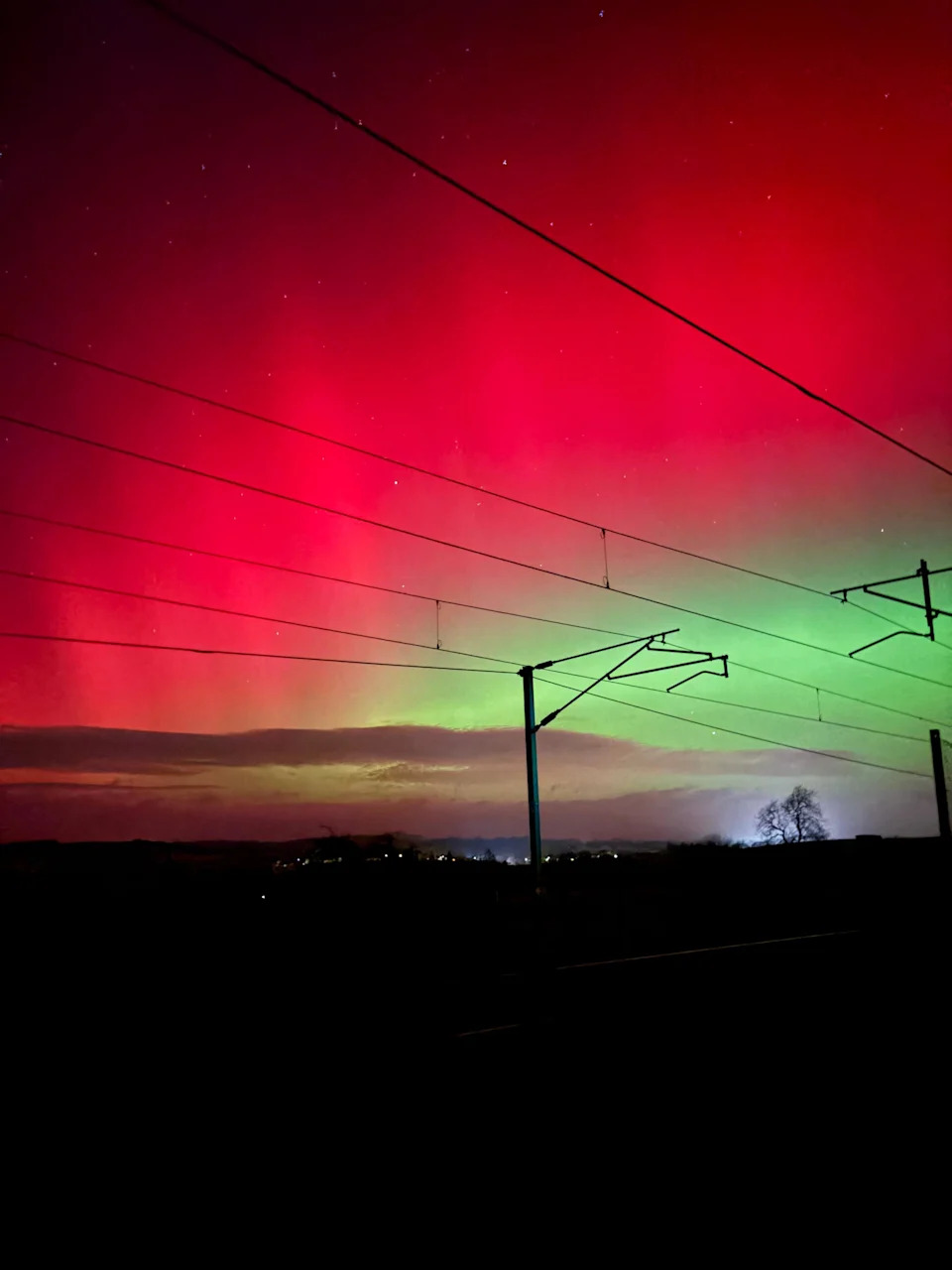 The aurora fills the sky with glowing red and green light above silhouetted railway power lines stretching across the scene.