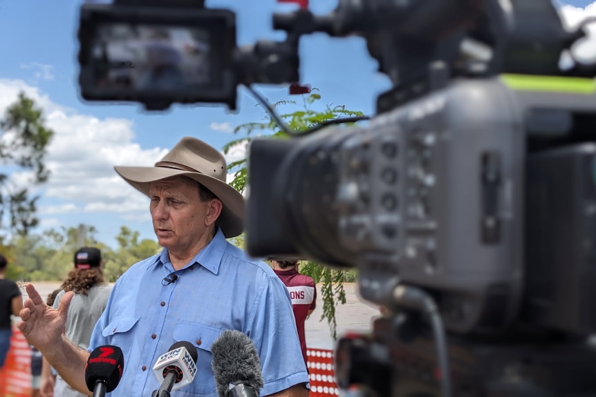 A man wearing a hat speaking in front of microphones behind a camera