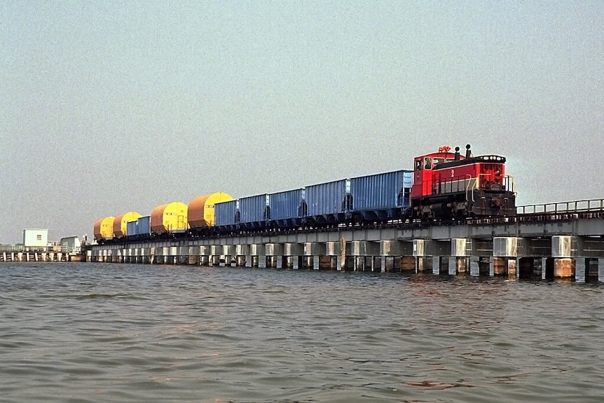 View across water of a red train engine hauling large cylindrical containers across a low bridge.