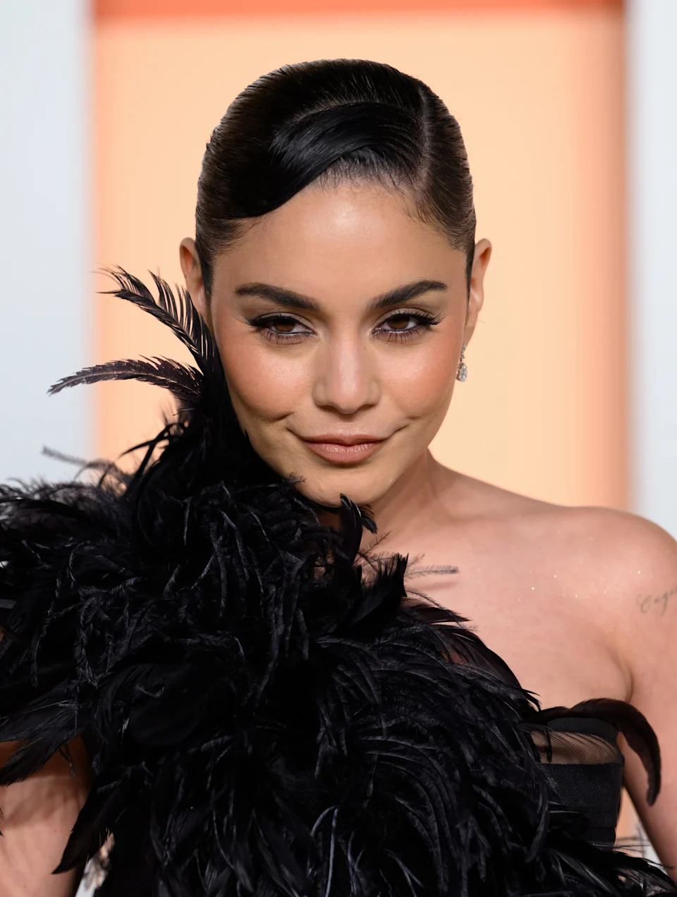 Vanessa Hudgens in elegant eathered outfit with slicked-back hair, posing confidently for a photo on a red carpet