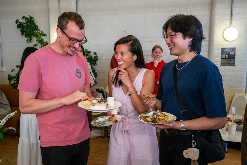 Three people with plates of food stand chatting and laughing