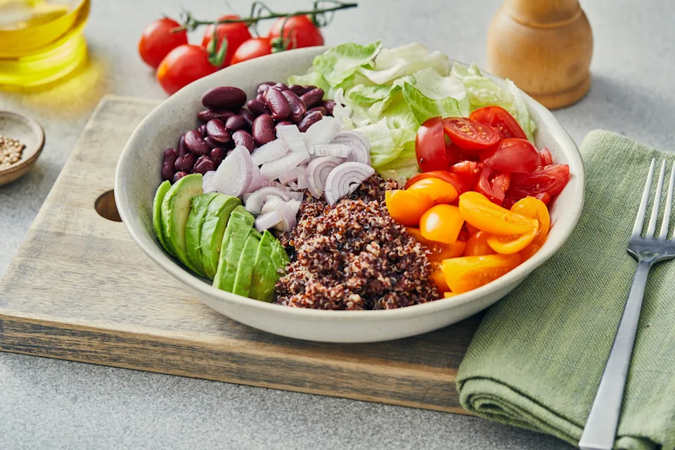 Bowl with quinoa, avocado, cherry tomatoes, kidney beans, sliced onion, and lettuce on a wooden board beside a green napkin and fork