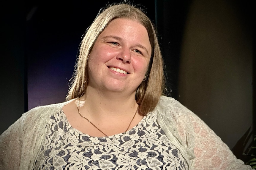 A woman with short hair and floral top smiles off to the side in a dark room.