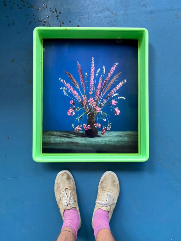 A person wearing white shoes and pink socks stands in front of a green tray holding a photo of a flower arrangement in a vase, set against a blue background on a wooden table.