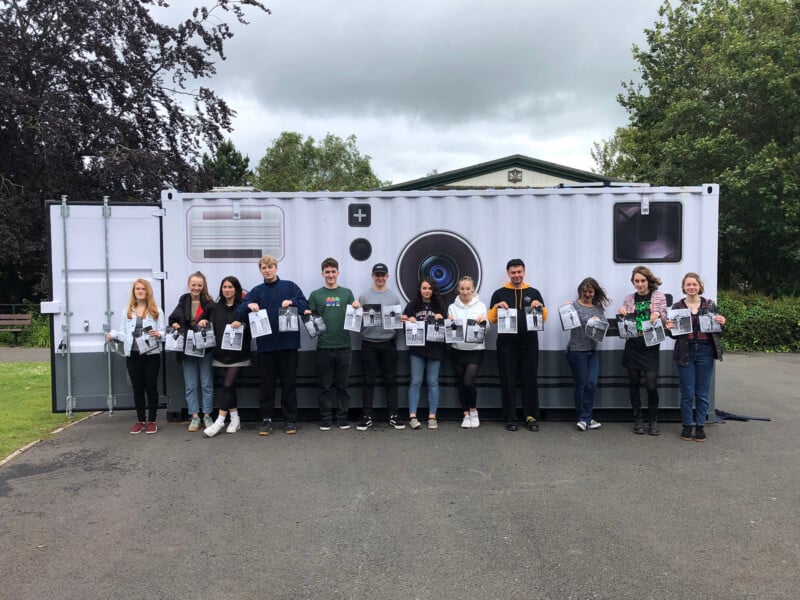 A group of thirteen people stands in a line outdoors, holding bags, in front of a white building decorated to look like a large camera. Trees and cloudy sky are visible in the background.