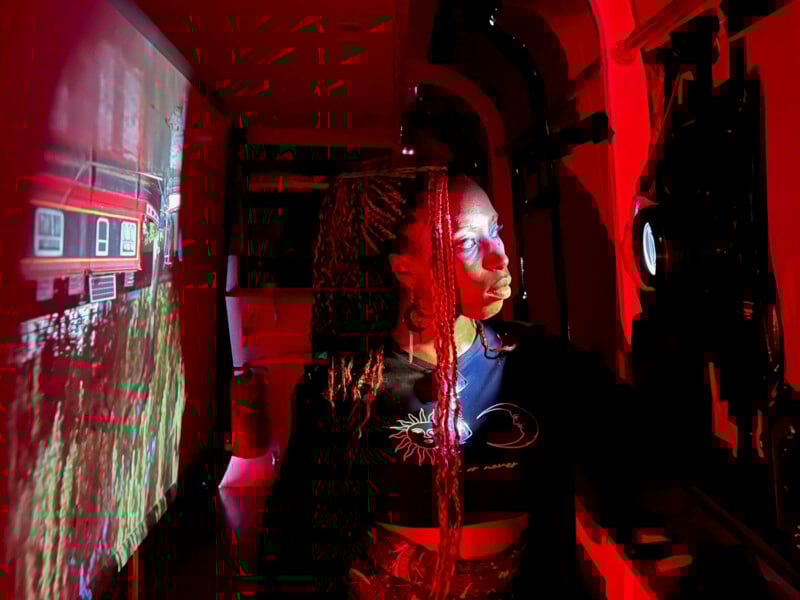 A woman illuminated by red light stands inside a room, facing a projector. A colorful image of a building and greenery is projected onto the wall beside her. Her face is partially lit by the projected light.