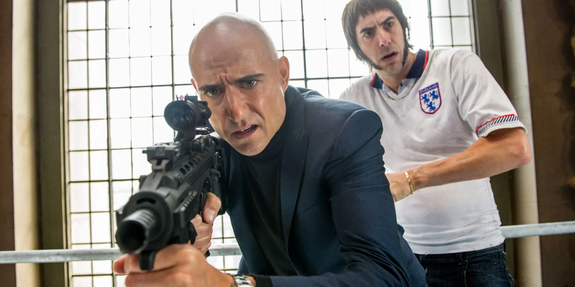 A man in suit holds a shotgun while another man in football jersey stands behind him