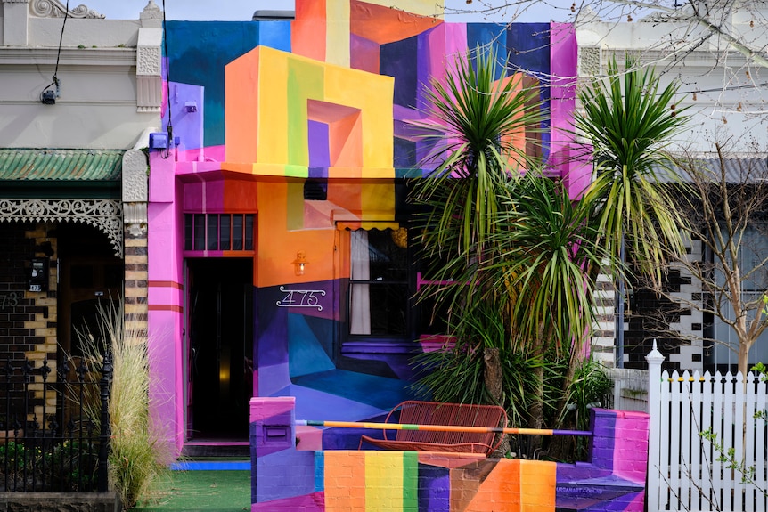 A terrace house with a vibrant multicolour mural as the facade is pictured with a towering yucca tree in front, from the street