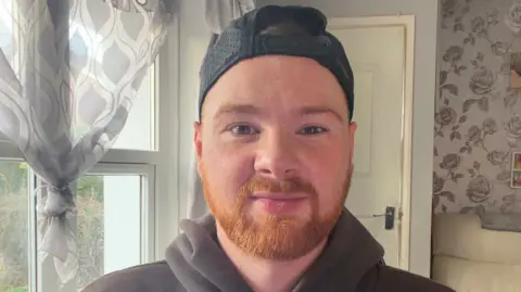 Ryan Jordan Ryan Jordan has a ginger beard and mustache. He is wearing a black cap backwards and a brown hoodie. He is stood in a living room. 