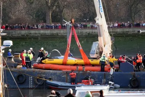 BDMLR A large crane is used to hoist the whale on to a large barge which has numerous people upon it. People are lined up behind a fence on the riverbank behind them