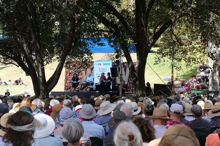 a large crowd of spectators sitting in a leafy Adelaide park watch four people talk on a stage at a festival.