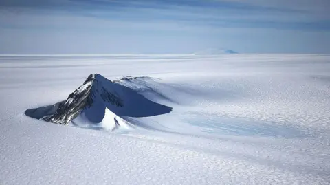 Getty Images A section of the West Antarctic Ice Sheet with mountain peaks poking out of the pristine, flat, white icy surface. The sky above is light blue with light clouds.