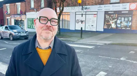 A man with a blade head, black rimmed glasses, a navy coat, checked shirt and yellow jumper. He is stood by a road with a zebra crossing. There is a darts shop at the other side of the road. Next to that shop, further down the road is a shop with two Union Jack flags outside called Booths. There is a silver car driving up the road.