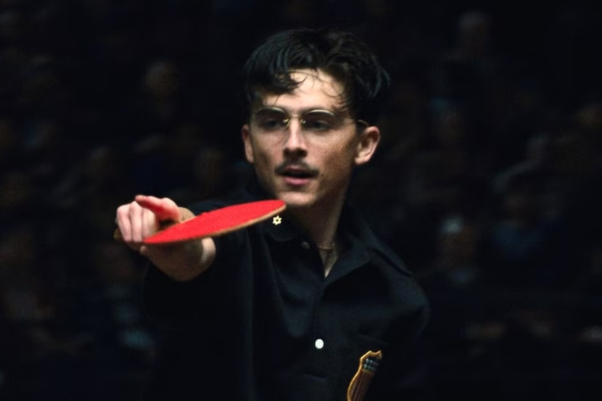 Timothee Chalament holds up a table tennis paddle and points with his index finger