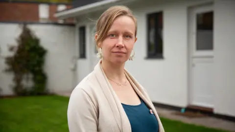 Dr Rosalind Baker-Frampton stood to the side with one of the Gordon Moody Gambling Charity offices behind her. She's stood outside on green grass and is looking directly at the camera. She's wearing a cream cardigan, with a blue t-shirt underneath and a name badge. She has light brown hair which she has tied in a bun.