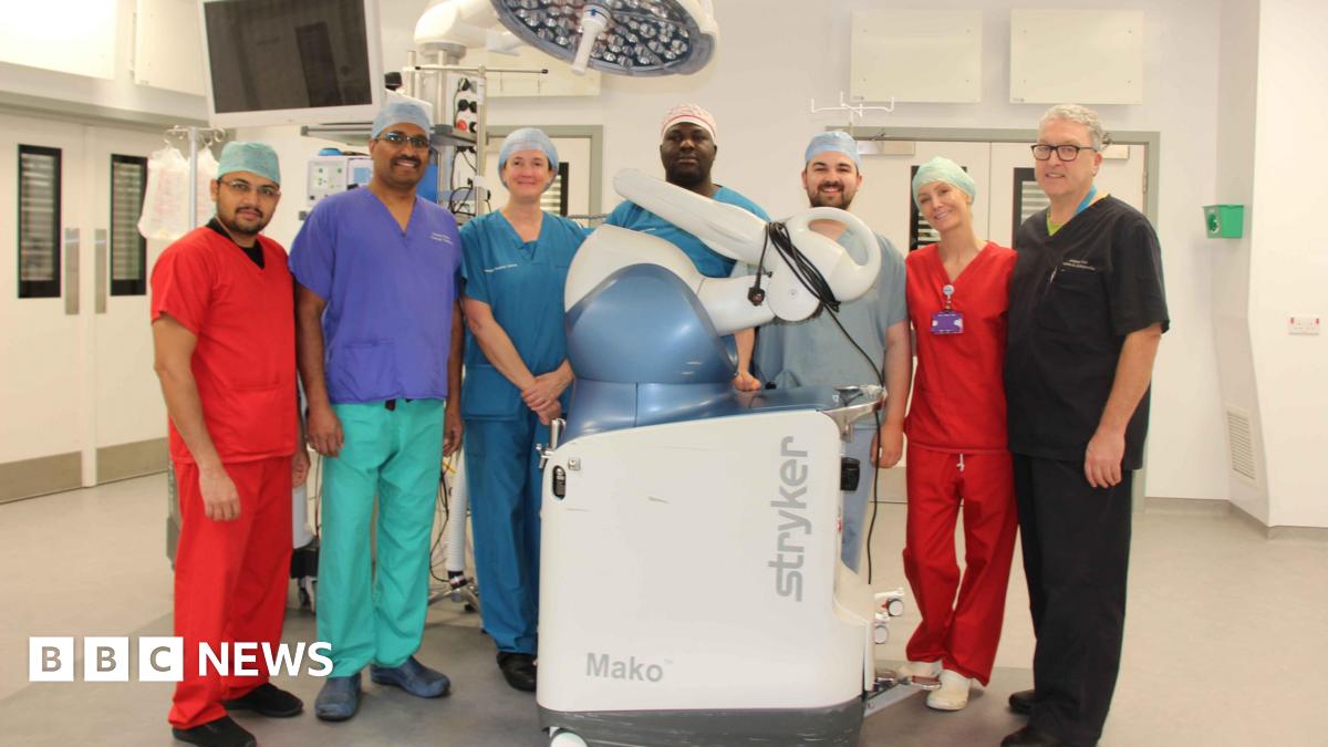 Andrew Port is a man with short white hair and glasses and is wearing dark scrubs. Next to him there are four woman and two men wearing scrubs of different colours. They are stood in a surgical room with the white and blue Mako robotic arm.