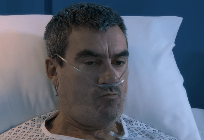 Cain Dingle in a hospital bed with tubes in his nose and an upset look on his face.