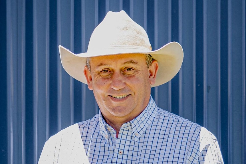 A man wearing a white hat and a blue and white check shirt smiles at camera.