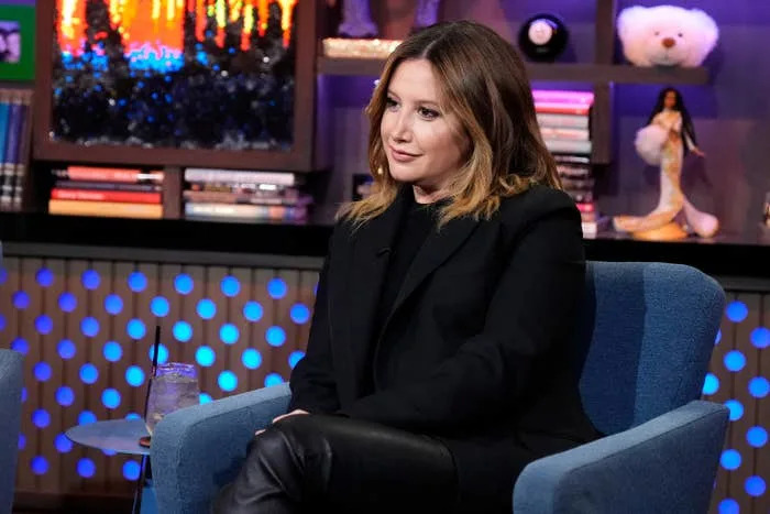 A person in a stylish black outfit sits in an interview setting with bookshelves and decorations in the background