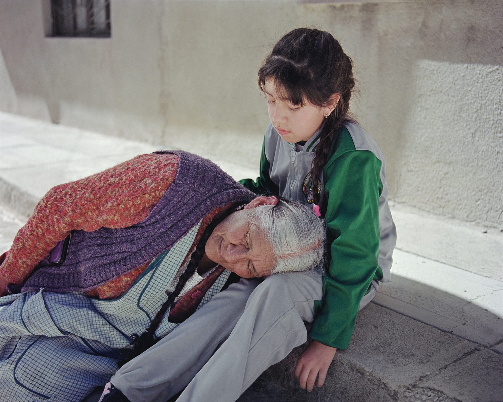 A photograph by River Claure of an elderly woman laying her head on the lap of a young girl