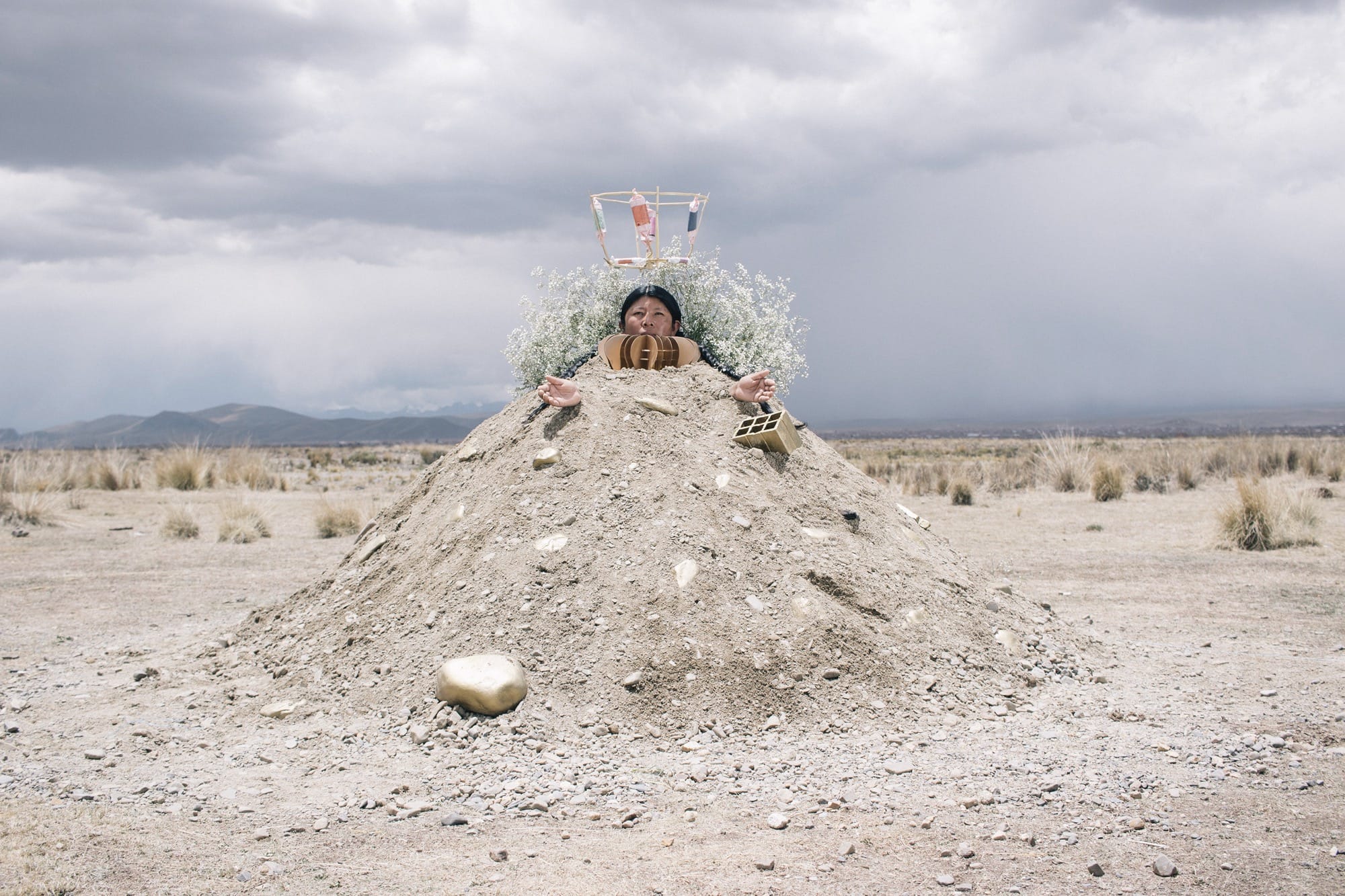 A photograph by River Claure of a figure inside of a mound of sand in the desert with a headdress, as if the sand is a gown