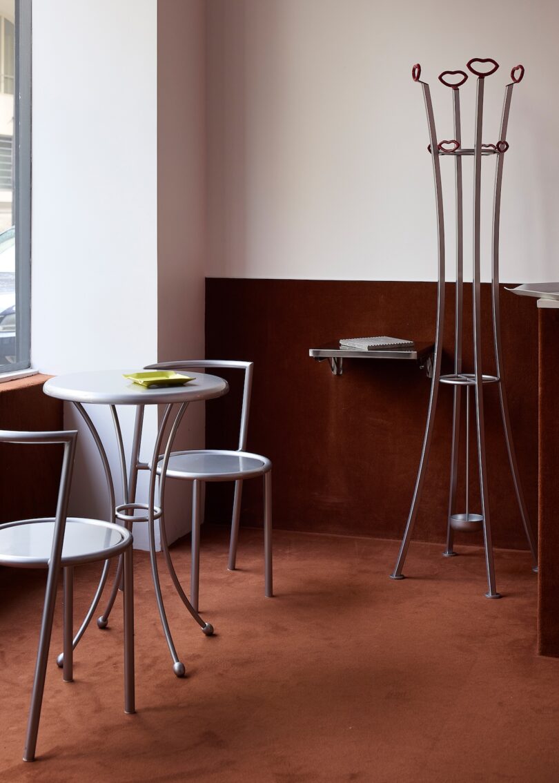 A small round table with two chairs, a wall-mounted foldable desk, and a tall coat rack in a room with brown carpet and white walls.