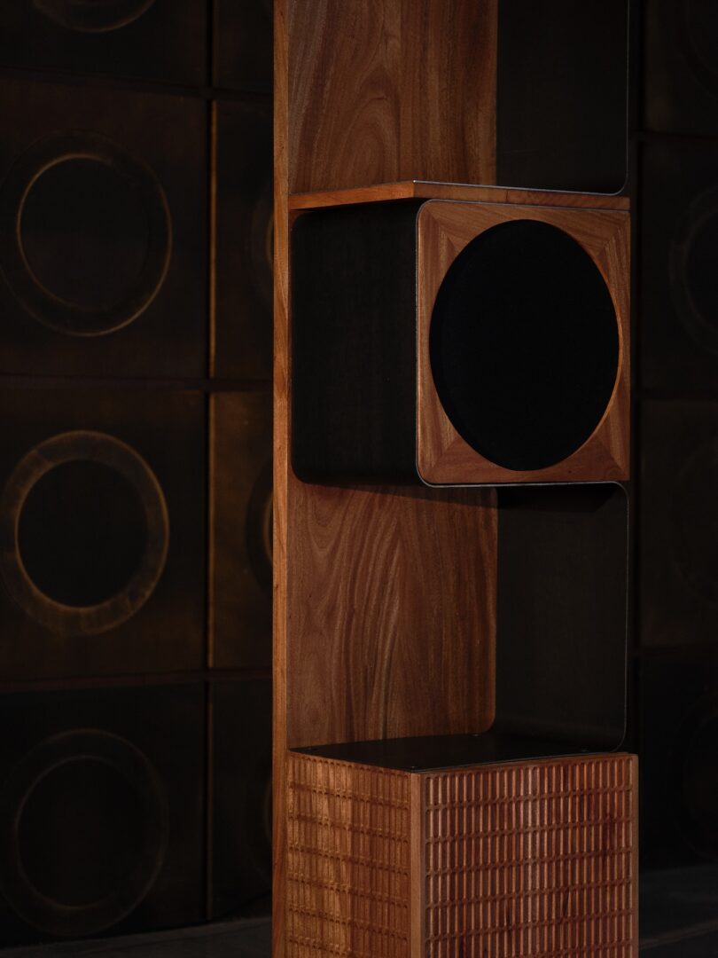 A modern wooden speaker with a circular black mesh front stands in front of a wall with a dark, circular pattern design.