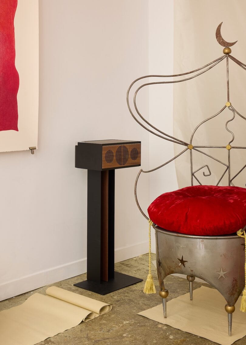 A metal chair with celestial designs and a red cushion is next to a black pedestal and a wall-mounted artwork; rolled papers are on the floor.