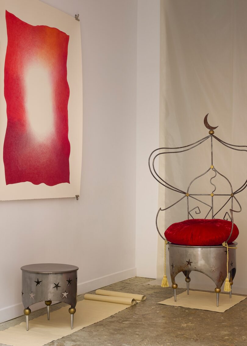 A metal chair with a red cushion and crescent moon design is beside a metal side table and an abstract red and white artwork on the wall in a minimalist room.