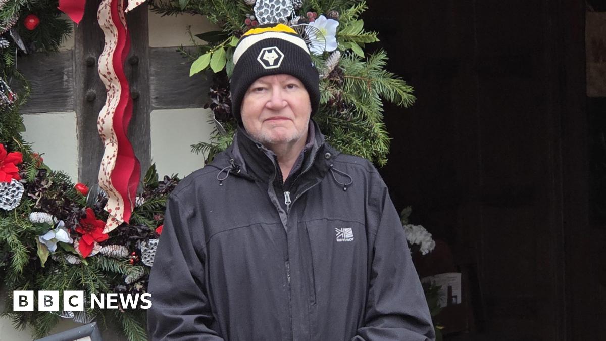 A man in a grey coat and yellow and black bobble hat in front of a wall with green plants and red ribbons