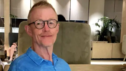 Steve Duell Head and shoulders portrait of Steve Duell, 65, wearing glasses and smiling gently. He is wearing a blue shirt and is in an area which could be a hotel or office lobby.