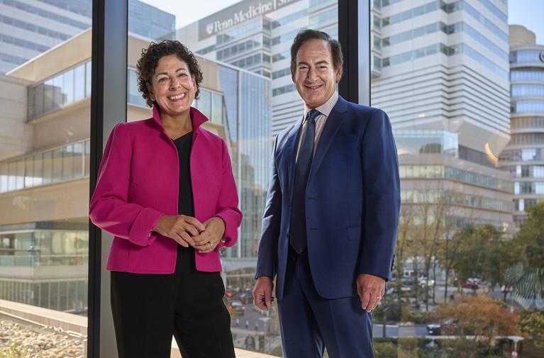 Researchers Angela DeMichele and Lewis Chodosh stand at the interior window of a large medical building.