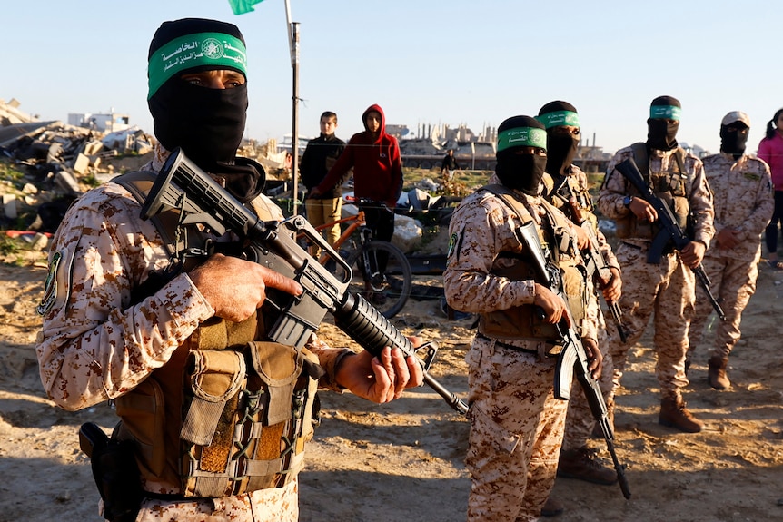 Men in military camo with black balaclavas, green headbands and assault rifles stand in a line