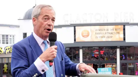 PA Nigel Farage stands in front of Clacton Pier with a microphone. You can see a "vote Reform" sign in the background and the amusements are behind him. He is wearing a blue suit and a pale blue tie, with a pale pink and blue checked shirt.